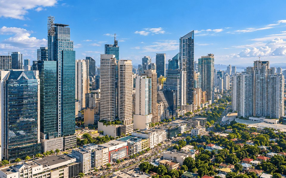 Manila skyline with glass towers, a busy tree-lined boulevard and residential rooftops under a bright blue sky