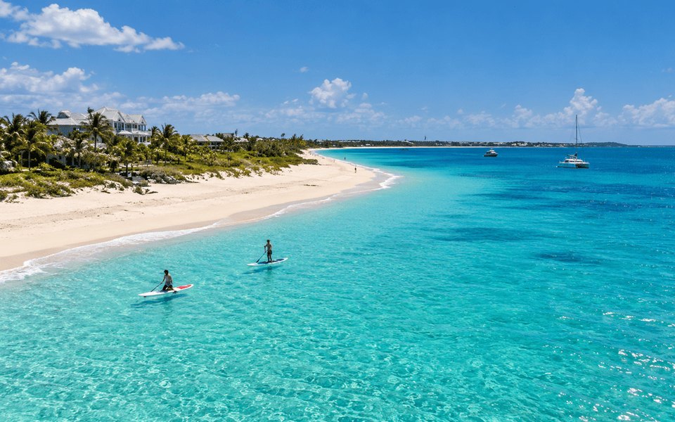 Two people paddleboarding in clear turquoise water off a white sand beach with luxury villas and palm trees in Providenciales, Turks and Caicos