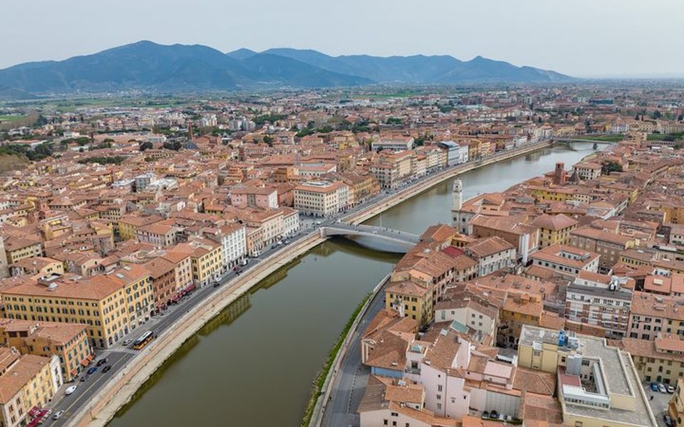 High-angle aerial view of the Arno River flowing through the historic centre of Pisa with terracotta rooftops and distant mountains