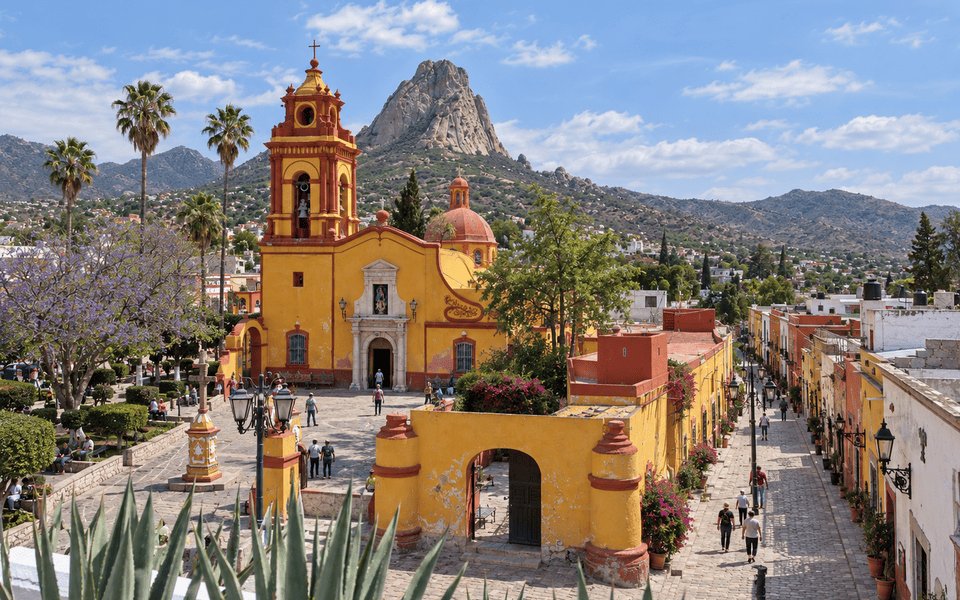 Yellow Parroquia de San Sebastián church in Bernal, Querétaro, with the Peña de Bernal monolith rising behind it under a clear blue sky